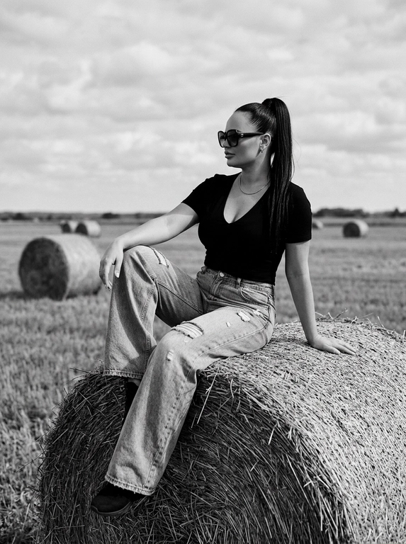 Чёрно-белая high-fashion editorial фотография в сельской местности, женщина с внешностью как на референсе, черты лица не изменять, композиция — более общий план (wide shot), камера на среднем расстоянии, фигура полностью в кадре с окружающим пространством, она сидит на большом тюке сена в поле, корпус слегка развернут, одна нога свободно опущена вниз, вторая согнута, создавая расслабленную, но выразительную позу, руки естественно лежат на колене и на поверхности тюка, взгляд направлен в сторону, профиль, выражение спокойное и уверенное, одежда: чёрный топ с V-образным вырезом и объёмные светлые широкие джинсы с потёртостями и рваными элементами, аксессуары: крупные солнцезащитные очки, минималистичные украшения, фон: открытое поле с несколькими тюками сена на разной глубине, создающими перспективу, линия горизонта ниже центра кадра, чтобы подчеркнуть небо, освещение: жёсткий естественный дневной свет с мягкими облаками, формирует выразительные тени и контраст, стиль: кинематографический, плёночное зерно, высокая детализация, реалистичная текстура кожи и ткани, редакционный fashion стиль, акцент на атмосфере и пространстве кадра


Какая будет прическа?: Гладкие волосы собраны в высокий хвост