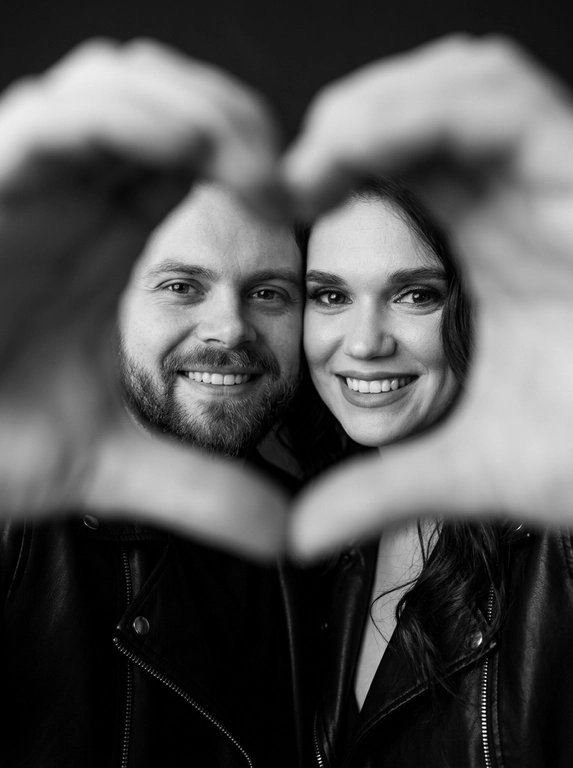 Ultra-realistic editorial photography of a smiling couple in a romantic and cinematic composition. The couple is centered in the background of the image, looking directly at the camera with a spontaneous, happy, and natural expression. In the foreground, extremely close to the lens and slightly out of focus, the couple’s hands form a heart shape perfectly framing their faces.

The composition creates strong visual depth, with sharp focus on the eyes and faces inside the heart formed by the hands. The hands occupy a large portion of the foreground, artistically blurred around the edges, creating a sophisticated cinematic effect.

Intimate, emotional, and elegant aesthetic inspired by modern perfume campaigns and premium lifestyle editorials. Soft studio lighting with delicate contrast and natural shadows. Minimalistic dark blurred background.

The couple wears minimalistic black leather jackets and sophisticated dark clothing. Genuine expressions, strong emotional connection, contemporary romantic atmosphere. Hyper-realistic skin texture, visible pores, detailed hair strands, authentic human appearance.

Black and white photography, rich tones, deep blacks, and soft highlights. 85mm lens, shallow depth of field, cinematic portrait photography, luxury editorial style, ultra detailed, hyper realistic, 8K.