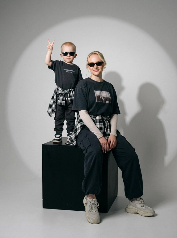 Photorealistic studio portrait photography in minimalistic fashion/editorial aesthetics, static camera at chest level of a seated person, light frontal angle without noticeable perspective deformation; composition with two characters: an model sits on a large matte black cube, body slightly turned towards the camera, knees bent, feet on the floor, the arms are lowered and relaxed in front of the knees, the fingers are neatly extended; the child stands on the same cube behind and to the left, at full height, one hand raised with a "V" gesture, the other lowered, pose confident and playful; both wearing narrow dark sunglasses, gaze directed at the camera; shooting plan — full height for both with a small supply of air above and below, center the center of the frame is shifted to the right to a sitting figure, a standing child balances on the left; the lens is normal/short portrait 50-70 mm, without width; aperture is approximately f/5.6–f/8, moderate depth of field, both characters are sharp, the background is slightly soft; the studio lighting is soft, with one key source on the front left at a level just above the head (softbox), giving smooth transitions and a pronounced but soft shadow against the background on the right; light filling light from the front reduces contrast, shadows are readable; the background is a smooth, light, seamless cyclorama/wall with a noticeable large oval light spot (gradient) in the center, there is a darker vignetting around, a clean minimalistic studio without props; the mood is calm, stylish, slightly retro, with an emphasis on street fashion and family vibe; the expression on the face of the seated model is a soft smile, relaxation; the person standing has a confident neutral facial expression; clothes: the seated model has a dark oversized T-shirt with a large rectangular print on the chest, under it a light longsleeve with long sleeves, wide dark cargo trousers/joggers with pleats at the knees and bottom, a plaid shirt/flannel tied at the waist with a knot, massive light sneakers with a pronounced sole and mesh texture; the child has a dark T-shirt with a large inscription on the chest, dark trousers, a plaid shirt tied at the waist, sneakers in two colors; fabrics with a realistic cotton and flannel texture, noticeable fine folds, natural creases on the knees and in the waist area, matte surfaces without gloss; micro-details: natural skin texture without plastic, neat glare on the lenses of glasses, a clear contour of the shadow from the raised hand on the background, clean floor and soft drop shadow from the cube; high detail, natural sharpness, light grain like a film shot, moderate contrast, soft matte/film look, no HDR, no overexposure and no unnecessary objects.

Какого цвета будут рубашки на поясе?: Чёрно-белый.