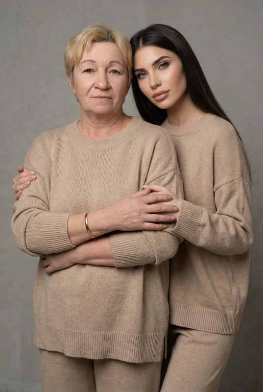 Используй эти два фото мамы и дочери и создай для них студийный портрет в чистом и элегантном стиле. Они стоят рядом, очень близко друг к другу. Дочь мягко обнимает маму за руку и слегка к ней прижимается со спины. Мама сложила руки перед грудью. Образ минималистичный, современный и стильный. На руке у матери тонкий золотой браслет. Профессиональный макияж и прическа.  Не менять черты лица ни на 1 процент. Фон — нейтральный серый с мягкой текстурой. Освещение рассеянное, мягкое, подчёркивающее естественные тона кожи. Атмосфера тёплая, нежная и спокойная, в стиле журнальной съёмки. Обе в свободных стильных бежевых свитерах и брюках.