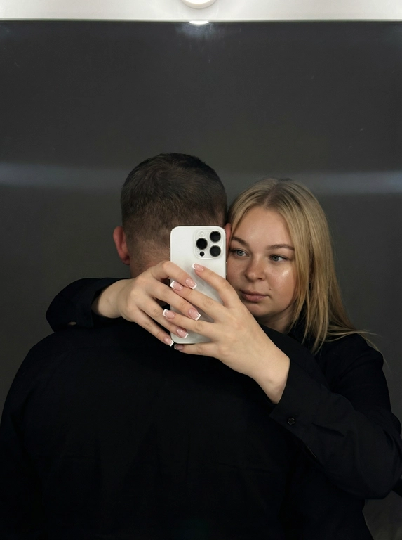 Ультрареалистичное кинематографичное зеркальное селфи пары. Строго сохранить лица из референса без изменений: возраст, пропорции, форму глаз, носа и губ, текстуру кожи, линию подбородка. Не изменять черты лица.

Локация: Серый лофт, low-key атмосфера.

Композиция: мужчина стоит спиной к камере, его лицо полностью не видно (back fully facing camera); за его спиной женщина обнимает его и снимает зеркальное селфи. Она держит белый смартфон Apple iPhone 16 Pro двумя руками. Фокус на руках и телефоне, френч квадратный маникюр. Физически корректное зеркальное отражение, без искажений..

Образ девушки: безупречный макияж, лёгкое сияние кожи.

Свет и стиль: подсветка от зеркала сверху-спереди, мягкие тени, тонкая горизонтальная световая линия на фоне. 50-85 мм, f/2-2.8, малая ГРИП; киноконтраст, плёночное зерно, high-end editorial, 8K.

Сцена полностью показана через отражение в зеркале, единственный источник изображения  — это отражение.

Ваша одежда: Мужчина и женщина одеты в чёрной рубашке;