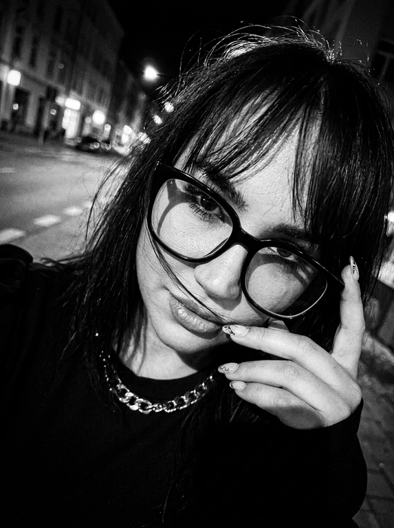 Интригующий, нуарный lifestyle момент; эстетика «messy chic» и ночного города; зернистое черно-белое фото с высоким контрастом, передающее атмосферу скрытности и дерзости.

Крупный план (extreme close-up), голландский угол (наклон камеры); имитация снимка на пленочную камеру 35mm с характерным цифровым шумом и виньеткой.

Модель игриво подносит руку к лицу, частично закрывая его пальцами; кончики пальцев касаются оправы очков или прядей волос.

Динамичный наклон головы в сторону; плечо слегка приподнято, создавая ощущение случайного, небрежного кадра «в моменте».

Загадочный, прямой взгляд в объектив сквозь линзы очков; губы слегка расслаблены; акцент на один глаз, видимый сквозь пряди волос.

Растрепанные волосы; тонкие пряди хаотично спадают на лицо и очки, создавая многослойность и текстуру.

Черный топ; акцент на аксессуары — крупные очки в массивной черной оправе.

Жесткий направленный свет (harsh flash) в темноте, создающий глубокие тени; эффект вспышки «в лоб», который выбеливает кожу и подчеркивает блеск очков и ногтей.

Выраженное пленочное зерно (film grain), легкий расфокус на заднем плане, естественные блики на линзах очков, детальная текстура кожи с сохранением естественных пор под слоем контрастных теней.

Photorealistic cinematic editorial, high-contrast black and white photography, raw aesthetic, 90s grunge vibe.  

Без: ИИ-эффектов, пластиковой кожи, пересветов, HDR, неестественных цветов. Все материалы (волосы, металл, ткань) выглядят максимально достоверно.