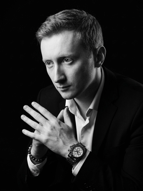 A hyper-realistic 8K cinematic portrait of a uploaded person,
captured in a dramatic Chiaroscuro style. The subject is seated at a
three-quarter angle, leaning slightly forward with a relaxed yet
commanding posture. His face is turned away from the lens, with
one side of his face clearly illuminated while the other is
completely lost in deep, velvety black shadows. His expression is
contemplative, not looking at the camera. The pose is precise: his
hands are clasped together near his chest; one hand is held
straight with fingers slightly tilted, while the other hand is
positioned so only the fingers are visible, resting over the first. He
wears a luxury black chronograph watch with a detailed metal link
bracelet, and a beaded bracelet. His attire is a sharp
black suit jacket over a white dress shirt with the top buttons
open, showing refined fabric textures. The background is a solid,
seamless black. The lighting is sharp and directional, highlighting
the intricate details of his hair,
the moisture in his eyes, and the glint off the watch face.
Cinematic black-and-white color grade with rich contrast and
smooth, natural skin transitions. Shot on 85mm lens, f/1.8, high-
resolution commercial photography