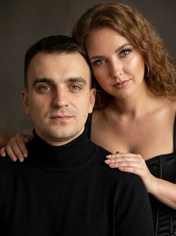 Гиперреалистичный студийный fashion-портрет молодой пары по загруженному референсу, 100% сходство лиц. Черты лица не изменять. Крупный план, кадр по плечи. Мужчина, спокойный уверенный взгляд прямо в камеру, естественное выражение лица, одет в черный трикотажный свитер с высоким воротом. Женщина стоит позади мужчины и нежно обнимает его, ее руки лежат на его плечах. Ее лицо расположено рядом с его лицом, она повернута слегка боком, голова немного наклонена вперед, взгляд направлен прямо в камеру. У женщины выразительные глаза с длинными густыми черными ресницами, естественный clean-glam макияж, натуральные брови, теплый оливковый оттенок кожи, небольшие серьги-кольца. Композиция плотная и интимная, их лица расположены очень близко друг к другу, современный модный портрет пары. Женщина одета в черный корсет. Кожа максимально натуральная: живая текстура кожи, видимые микропоры, subtle skin glow, естественный мягкий блеск кожи, natural skin microtexture, skin translucency, micro skin details, легкие естественные несовершенства кожи, без ретуши и без эффекта пластика. Освещение: мягкий студийный beauty lighting, рассеянный ключевой свет спереди и немного сбоку, мягкие глубокие тени для объема лица, деликатные световые блики на коже. Фон: глубокий темный студийный фон, темно-графитовый или почти черный, создающий контраст с кожей и лицами. Стиль: luxury fashion editorial photography, high fashion portrait, ultra photorealistic, 85mm portrait lens, shallow depth of field, creamy bokeh, cinematic soft lighting, RAW photo, extremely detailed, fine hair strands, natural skin pores, professional photography, photorealism, 8k, high dynamic range.