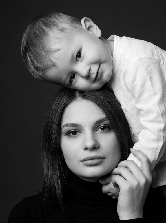 Родитель и ребёнок с внешностью как на приложенных фото, художественная семейная студийная фотосессия. Композиция — вертикальный портрет, крупный план лиц. Родитель внизу смотрит прямо в камеру, лицо фронтально. Ребёнок сверху, голова повернута набок, щека лежит на голове родителя, смотрит в камеру с нежной улыбкой. Рука ребёнка обнимает родителя за шею, видна в кадре, родитель держит руку ребёнка своей рукой.
Одежда: ребёнок в белой льняной рубашке, родитель в чёрной водолазке. 
Фон — тёмно-серый почти чёрный однотонный студийный, создаёт контраст. Освещение — мягкое боковое студийное, создаёт объём и глубину.
Чёрно-белая художественная фотография с выразительными контрастами. Атмосфера нежная, интимная, спокойная.