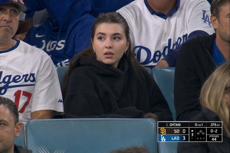 Person sitting in the crowd at a professional baseball game (or it could be any other match that I will write below), filmed from far away by a live broadcast camera. 
Seated in blue stadium chairs, slightly leaned back, looking off to the side with a surprised caught-on-camera expression, lips slightly parted.

Какая будет одежда? : Оверсайз худи черного цвета и джинсы