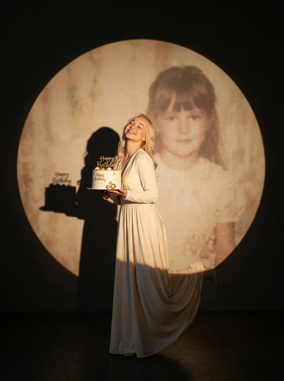 Create ultra-realistic cinematic birthday photoshoot, elegant studio with a dark background and a large circular spotlight projection on the wall. Inside the spotlight, a faded vintage childhood photo of the same girl(the reference image 2nd given)appears as a nostalgic memory, slightly transparent with old photo texture and soft blur. In the foreground, the present-day version of the same model (the reference image 1st given)  holding a white birthday cake decorated with gold accents with a “Happy Birthday". 

If the model is a woman, then she is wearing a cream long gown. If the model is a man, he is wearing a suit.

her head tilted back, eyes closed, natural smile,  cinematic shadow cast on the wall, warm soft spotlight lighting.

If the model was a woman, she additionally lifted her leg in an elegant pose.