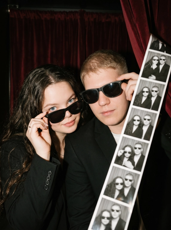 Крупный план (close-up) эффектной пары — женщины и  мужчины (не меняй лица), позирующих внутри тесной винтажной фотокабины. Они сидят вплотную друг к другу, создавая атмосферу ночного города, свидания или светской вечеринки. Композиция плотная и динамичная.

Одежда и стиль:
*   Женщина (слева): В классическом черном пиджаке. Одной рукой она кокетливо поправляет свои черные солнцезащитные очки. У нее безупречный вечерний макияж с влажным блеском на губах.
*   Мужчина (справа): Он одет в строгий черный пиджак, на нем также надеты классические черные солнцезащитные очки (Wayfarer или авиаторы), которые он слегка придерживает рукой.
*   Образ пары выдержан в едином стиле «Total Black» и эстетике «Old Money» или «Secret Agent».

Окружение и детали:
Действие происходит внутри аутентичной фотокабины. Справа на переднем плане видна свисающая длинная лента готовых черно-белых фотоснимков (photobooth film strip), на которых запечатлена эта же пара в разных позах. Фон темный, с тяжелыми складками занавески, что подчеркивает интимность момента.

Стиль и освещение:
Освещение имитирует резкую вспышку фотокабины — яркий фронтальный свет, создающий глубокие тени и подчеркивающий текстуру кожи и блеск аксессуаров. Цветовая палитра приглушенная, монохромная с теплыми телесными оттенками. Атмосфера «Nightlife Luxury».

Высочайшая детализация, акцент на блики в очках и текстуру ткани пиджаков, реалистичные черты лиц, кинематографичное качество, гиперреализм, 8k.