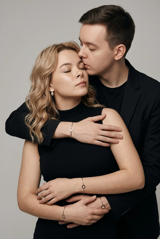 Мужчина и женщина позируют в студии в объятиях. Мужчина стоит позади женщины, обнимая её обеими руками; его тело повернуто к ней, голова слегка наклонена вниз, губы касаются её лба. Женщина немного отклоняет голову назад к его плечу, её глаза закрыты. Кадр — средний, от талии до головы, снят на уровне глаз. Композиция плотная, акцентирует линии рук и соприкосновение лиц. Женщина стоит, прямо обращённая к камере.
На ней облегающий чёрный топ без рукавов с высоким воротом, из плотной матовой ткани.
Мужчина одет в чёрную футболку и чёрную неброскую куртку. На обоих запястьях — тонкие металлические браслеты с минималистичными круглыми подвесками, а женщина носит небольшие светлые металлические серьги с гладкой поверхностью.
Обстановка минималистичная: однотонный светло-серый фон без текстуры.
Освещение — мягкий студийный свет: рассеянный фронтальный и боковой свет создаёт плавные переходы света и тени на лицах и руках. Тени мягкие, средней глубины. Контраст умеренный, без жёстких бликов. Свет равномерно распределён по фигурам, фон подсвечен мягко, без выраженных градиентов.
Съёмка на портретный объектив 85 мм, малая глубина резкости с чётким фокусом на лицах и руках; фон слегка размыт, текстуры кожи и ткани высоко детализированы. Это минималистичный студийный портрет, высокодетализированный и фотореалистичный.
Минималистичное романтическое студийное настроение, нейтральные тёплые тона кожи, низкая–средняя насыщенность, мягкое рассеянное освещение, средний контраст с нежными тенями, плавные переходы бликов, естественная передача кожи, чистый светлый фон, тонкий тональный баланс, современная мягкая редакционная цветокоррекция.

Волосы женщины с укладкой по плечи с аккуратно уложенными прядями.