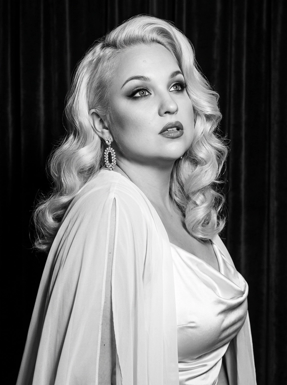 Фотореалистичный контрастный чёрно-белый glamour/editorial портрет в стиле Old Hollywood, вертикальный кадр по грудь. Женщина в 3/4, корпус чуть наклонён вперёд; голова повернута влево, взгляд вверх/в сторону (не в камеру), губы чуть приоткрыты, выражение драматичное и кинематографичное. Образ: белая накидка из шифона с длинными струящимися рукавами, под накидкой драпированный атласный белый топ; на ушах крупные серьги со стразами/каскадом. Макияж: классический вечерний glam (в ч/б читается как контраст) — чёткие брови, мягкие smoky тени, тонкая стрелка, длинные ресницы, губы с чётким контуром и сатиновым финишем. Фон: тёмный студийный с мягким рисунком/драпировкой (как бархатные шторы), размытый. Свет: жёсткий key слева спереди (beauty dish) + контровой/rim справа для отделения волос, глубокие тени, яркие блики на скулах и волосах, богатый тональный диапазон (глубокие чёрные и сливочные белые). Камера: 85mm, f/2.0–2.8, ISO 100–200, 1/160, резкость на глазах и губах, фон в мягком боке, лёгкое плёночное зерно, без “пластика”.

Какая будет укладка? : Волосы: объёмные голливудские волны, аккуратная укладка с сильным объёмом и мягкими завитками, блеск волос.