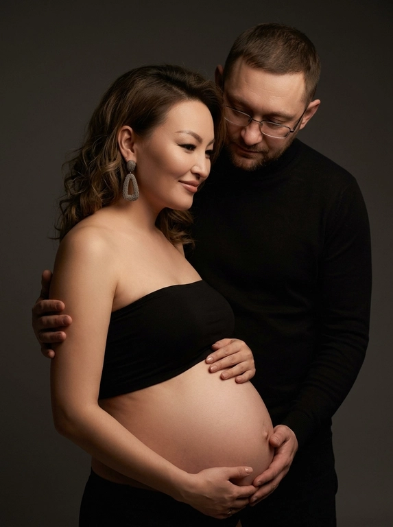 Интимная стильная сцена с трогательной эмоциональной связью. Женщина в полупрофиле к камере с легкой улыбкой, в минималистичном черном топе без бретелек, показывающий открытый беременный живот с мягкой матовой кожей под равномерным светом, руки на животе, создавая заботу. Крупные висячие серьги с видимой фактурой и блеском. Мужчина позади, наклонив голову вниз к ней, в черном тонком свитере с высоким воротом, подчеркивающем стройность. Мягкое освещение, серая фональная поверхность, тени подчеркивают рельефы и создают игру света и тени, как тихий вечер в закрытом пространстве. Композиция: мужчина словно обнимает женщину, руки частично обрезаны. Лицевые черты сохранены, передавая индивидуальность и естественную красоту, атмосфера любви, ожидания и легкой загадочности. Фотореалистично, высокая текстурная детализация, 8K.