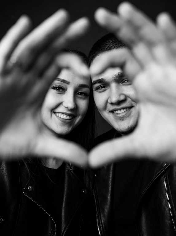 Ultra-realistic editorial photography of a smiling couple in a romantic and cinematic composition. The couple is centered in the background of the image, looking directly at the camera with a spontaneous, happy, and natural expression. In the foreground, extremely close to the lens and slightly out of focus, the couple’s hands form a heart shape perfectly framing their faces.

The composition creates strong visual depth, with sharp focus on the eyes and faces inside the heart formed by the hands. The hands occupy a large portion of the foreground, artistically blurred around the edges, creating a sophisticated cinematic effect.

Intimate, emotional, and elegant aesthetic inspired by modern perfume campaigns and premium lifestyle editorials. Soft studio lighting with delicate contrast and natural shadows. Minimalistic dark blurred background.

The couple wears minimalistic black leather jackets and sophisticated dark clothing. Genuine expressions, strong emotional connection, contemporary romantic atmosphere. Hyper-realistic skin texture, visible pores, detailed hair strands, authentic human appearance.

Black and white photography, rich tones, deep blacks, and soft highlights. 85mm lens, shallow depth of field, cinematic portrait photography, luxury editorial style, ultra detailed, hyper realistic, 8K.