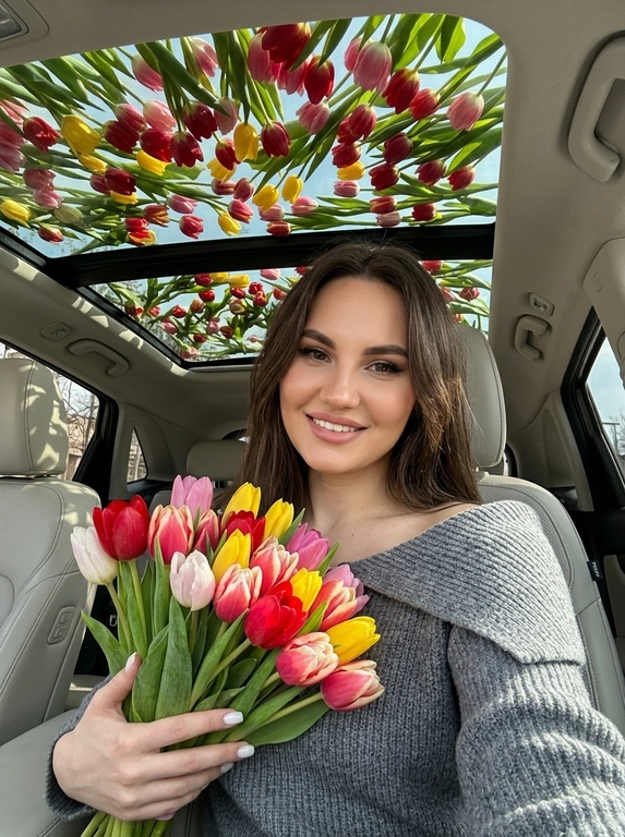 Ультрареалистичная фотография женщины, сидящей в современном автомобиле с большой панорамной стеклянной крышей. На коротких ногтях маникюр молочного цвета. Она сидит, делая селфи и держит букет тюльпанов (разные цвета). Естественный макияж.

Над панорамной крышей раскинулся густой полог из тюльпанов (разных цветов и сортов), покрывающий все небо. Тюльпаны хаотично лежат на всей поверхности стеклянной крыше (обязательно лежали на поверхности). Мягкий естественный солнечный свет пробивается сквозь тюльпаны, создавая красивые тени внутри автомобиля. Кинематографическое освещение, высокая детализация, роскошная лайфстайл-фотография, 8K. Не меняй пол, возраст, внешность, цвет волос.

Какой будет образ на фото?: На ней серый вязаный джемпер с широким отворотом, который носится открытыми плечами.
