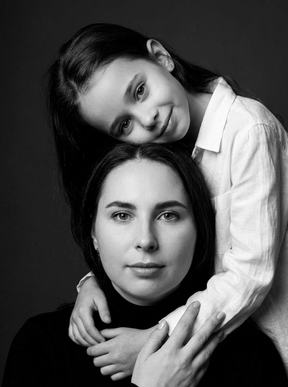 Родитель и ребёнок с внешностью как на приложенных фото, художественная семейная студийная фотосессия. Композиция — вертикальный портрет, крупный план лиц. Родитель внизу смотрит прямо в камеру, лицо фронтально. Ребёнок сверху, голова повернута набок, щека лежит на голове родителя, смотрит в камеру с нежной улыбкой. Рука ребёнка обнимает родителя за шею, видна в кадре, родитель держит руку ребёнка своей рукой.
Одежда: ребёнок в белой льняной рубашке, родитель в чёрной водолазке. 
Фон — тёмно-серый почти чёрный однотонный студийный, создаёт контраст. Освещение — мягкое боковое студийное, создаёт объём и глубину.
Чёрно-белая художественная фотография с выразительными контрастами. Атмосфера нежная, интимная, спокойная.