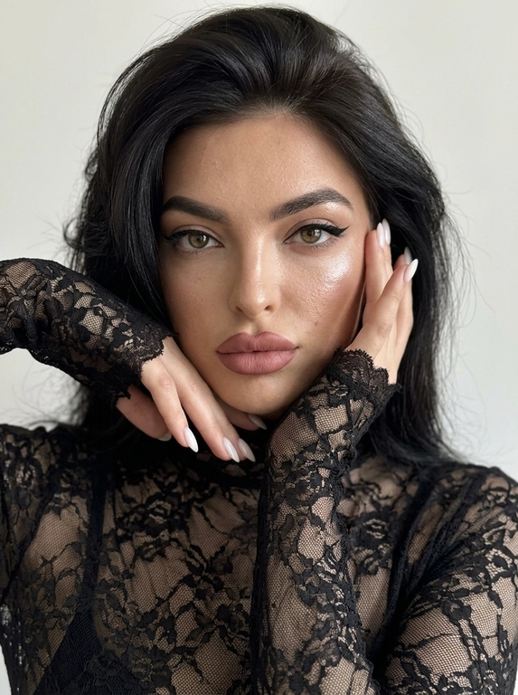 Лёгкий макияж с акцентом на глаза: чёрные cat-eye стрелки на верхнем веке, нюдовая матовая помада оттенка пыльной розы, очерченная карандашом для губ на тон темнее, влажный хайлайтер. Кожа загорелая. Ультрареалистичная фотография, естественная текстура кожи, лёгкий шум, необработанный реалистичный вид, без фильтра красоты, без CGI, visible eye capillaries, natural skin texture with pores and micro-imperfections, depth of field, subsurface scattering, soft morning light, Tyndall effect, hyper-detailed iris, unedited RAW photo, 8K resolution. Максимально фотореалистичный студийный портрет, вертикальный формат, снято на iPhone 17 Pro, 8K.

Женщина сидит, но кадр настолько близкий, что место, на котором она сидит, бёдра и ноги полностью остаются за пределами кадра. РАКУРС ОЧЕНЬ БЛИЗКИЙ: камера прямо перед женщиной, на уровне глаз, объектив близко к лицу. В кадре только голова, плечи, руки и небольшая часть верхней части корпуса. Макушка близко к верхней границе кадра. Лицо почти фронтально, совсем немного повёрнуто в сторону, взгляд прямо в объектив, губы слегка приоткрыты. Женщина сидит расслабленно, корпус слегка подан вперёд.

Обе руки подняты к лицу очень компактно. Локти находятся близко друг к другу перед корпусом и направлены вниз, не разведены в стороны. Одна кисть поднята снизу к подбородку и губам, пальцы мягко согнуты, указательный палец касается нижней губы. Вторая кисть находится немного выше, возле противоположной щеки. Запястья практически рядом. Руки располагаются по обе стороны лица, возле щёк и висков.

На женщине тонкая чёрная полупрозрачная кружевная кофта по фигуре с высоким облегающим воротником. Всё полотно состоит из нежного ажурного кружева с мелким органичным растительным рисунком из тонких переплетающихся стеблей, небольших цветов и листьев, между которыми просвечивает кожа. Это цельное тонкое кружевное полотно, не сетка с нашитой вышивкой и не тяжёлый гипюр. Рукава экстремально длинные, закрывают запястья и почти полностью заходят на кисти до оснований пальцев. Края рукавов необработанные: кружево заканчивается непосредственно по своему рисунку, образуя тонкие ажурные выступы вокруг кистей, без манжет, подгиба и ровной строчки.

Макияж выразительный: загорелая, увлажнённая, сияющая кожа с натуральной текстурой и порами, сильный бронзер и контуринг скул, влажный хайлайтер, вытянутые чёрные кошачьи стрелки, пухлые нюдово-розовые губы с более тёмным коричнево-розовым контуром.

Позади неё ровная однотонная молочная стена: светлый мягкий молочно-белый оттенок, не серая, не серо-молочная, не бежевая, без декора и предметов. Мягкий нейтральный студийный свет, натуральный баланс белого, естественные оттенки кожи. Никакой жёлтой, золотистой или тёплой обработки. Чистая натуральная фотография без фильтров, пресетов, цветокоррекции, винтажного эффекта, HDR, ретуши, боке, пластиковой кожи и ИИ-эффекта. Маникюр молочный, миндальной формы, ровный и аккуратный.

Причёска: Волосы с сильным воздушным объёмом у корней, уложены на крупный брашинг. Передние объёмные слои проходят вдоль висков и щёк, слегка приминаются поднятыми руками и ниже снова становятся пышными. Несколько тонких прядей естественно проходят возле щёк и губ. Никакой расчёски, щётки или других предметов в кадре.  

Доп. характеристики: Не улыбаться