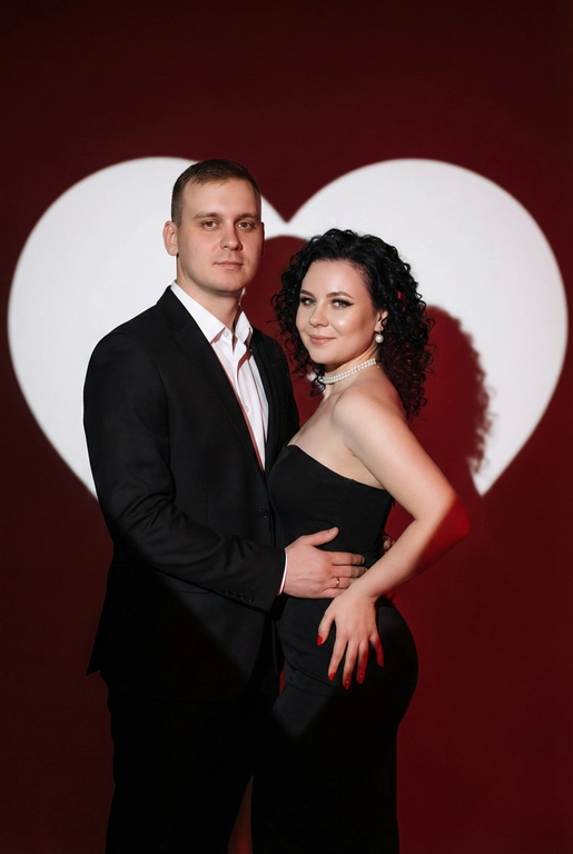 Фотореалистичный редакционный крупный портрет по пояс для пары по референсным фото. Мужчина и женщина позируют вместе, стоят близко. Мужчина одной рукой приобнимает женщину за талию. Фон: тёмно-красный студийный задник, большое белое сердце, проецируемое на стену. Сохранить внешность обоих: реальные черты лица, естественная текстура кожи, реальный цвет и длина волос, без ретуши и изменений. Женщина: облегающее чёрное вечернее платье без бретелей с небольшим разрезом, жемчужный чокер, жемчужные серьги, красный маникюр. Поза в три четверти, спина слегка выгнута, одна рука на поясе или касается руки мужчины. Лёгкая улыбка, взгляд в камеру. Мужчина: чёрный классический костюм, белая рубашка. Прямая осанка, уверенная поза. Взгляд в камеру, легкая улыбка. Свет: драматическое боковое студийное освещение, высокий контраст, чёткие тени на фоне. Композиция: вертикальный кадр по пояс, пара в центре, негативное пространство вокруг сердца. Стиль: фотореализм, модная редакционная съёмка, кинематографическая цветокоррекция (тёплые красные, глубокие чёрные), высокая детализация. Не менять мою прическу.