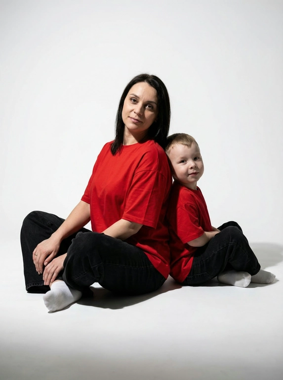 Гиперреалистичная фотография. Качество 8К. Используя портретное сходство с людьми с фотографий, создай фотографию: женщина в красной оверсайз свободной футболке и свободных черных джинсах, белых носках. Ребенок одет в красную оверсайз футболку, черные свободные джинсы и белые носочки. Женщина сидит на полу, согнув колени, смотрит прямо в кадр с еле заметной улыбкой. Сзади женщины в такой же позе сидит ребенок облокотившись спиной к спине женщины, с легкой улыбкой. Темный стиль модного редакционного журнала, глубокие тени и драматическое студийное освещение, белый фон, элегантная и мощная композиция, ультрареалистичный, высокая детализация, художественный портрет.