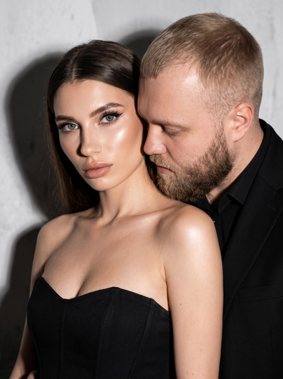 Не меняя черты лица, студийная fashion-съемка пары в минималистичном интерьере с серо-белым фоном, атмосфера гламура и интимной близости, ракурс — крупный портрет по грудь с легким поворотом корпуса, женщина на переднем плане, мужчина позади склоняется к её плечу, поза — она слегка отвернута от камеры с прямым взглядом, он касается лицом её шеи/плеча, одежда — на женщине черный корсет/бандо с открытыми плечами, на мужчине классический черный пиджак и рубашка, макияж — glam (ровный сияющий тон, сильный хайлайтер, четкий контуринг, стрелки, густые ресницы, глянцевые губы), свет — жесткая фронтальная вспышка + мягкий заполняющий свет, выраженные тени позади, эффект flash photography / editorial, холодно-нейтральная цветокоррекция с контрастом и глянцем кожи, высокая детализация, slight film grain, shallow depth of field, съемка на Canon EOS R / 50mm, f/2.0, direct flash, studio look.