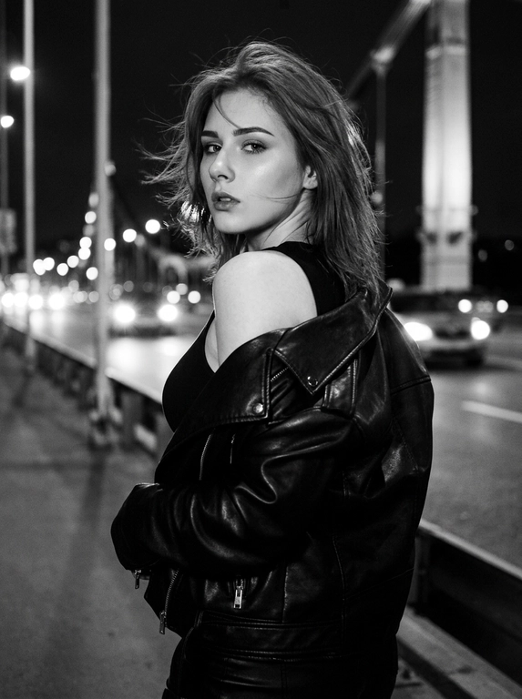 Используй внешность модели с фото. Фотореалистичное черно-белое фото в стиле модной редакционной съемки с эстетикой пленочной фотографии (зернистость, высокий контраст). Модель с волосами слегка растрепанными ветром. Она одета в черную, объемную кожаную куртку-косуху, которая небрежно спущена с левого плеча, открывая его. Под курткой надет черный облегающий топ. Поза: модель стоит вполоборота спиной к камере, но резко оборачивается и смотрит назад через плечо прямо в объектив. Выражение лица серьезное, дерзкое и чувственное, губы слегка приоткрыты, густые ресницы. Фон размыт, видны элементы конструкции моста и огни проезжающих машин вдали, создающие городскую атмосферу. Освещение естественное, подчеркивающее текстуру кожи и блеск кожи куртки. Высокое разрешение, 8k. Освещение мягкое и равномерное, без жестких теней, свет подчеркивает блеск жемчуга и страз, цвета насыщенные, но реалистичные. Высокая детализация, фотореализм, четкий фокус на лице, естественные пропорции.