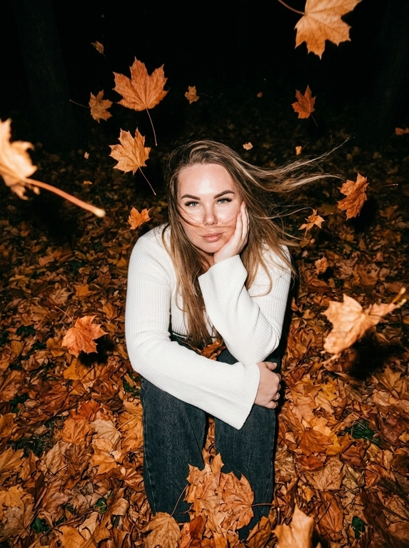 Ultra-realistic candid NIGHT autumn photo. The woman sits curled up directly in a thick carpet of dry autumn maple leaves, knees pulled toward her chest. Camera positioned very high above her, almost directly overhead, dramatic top-down perspective. She raises her face toward the camera, one hand gently supporting her cheek, the other arm resting across her knees.
She wears a fitted white knitted top with very long dramatic FLARED BELL SLEEVES, fitted through the arms and strongly widening from the elbows.
Strong autumn wind blows her hair.

It is DARK outside, almost nighttime. The background beyond the flash falls into deep natural darkness. Shot with a powerful direct on-camera flash: her face, white sweater and the closest leaves are brightly illuminated, while distant leaves rapidly disappear into black shadows. Dozens of dry orange, rust and brown leaves are FLYING THROUGH THE AIR around her, caught mid-flight by the flash; some pass very close to the lens and create strong realistic motion blur and streaks. A few strands of hair also have motion blur from the wind. Face remains sharp. Raw spontaneous flash photography, high contrast, deep blacks, warm saturated autumn tones, slight grain, imperfect handheld framing, realistic textures, no studio lighting, no HDR, no AI look.