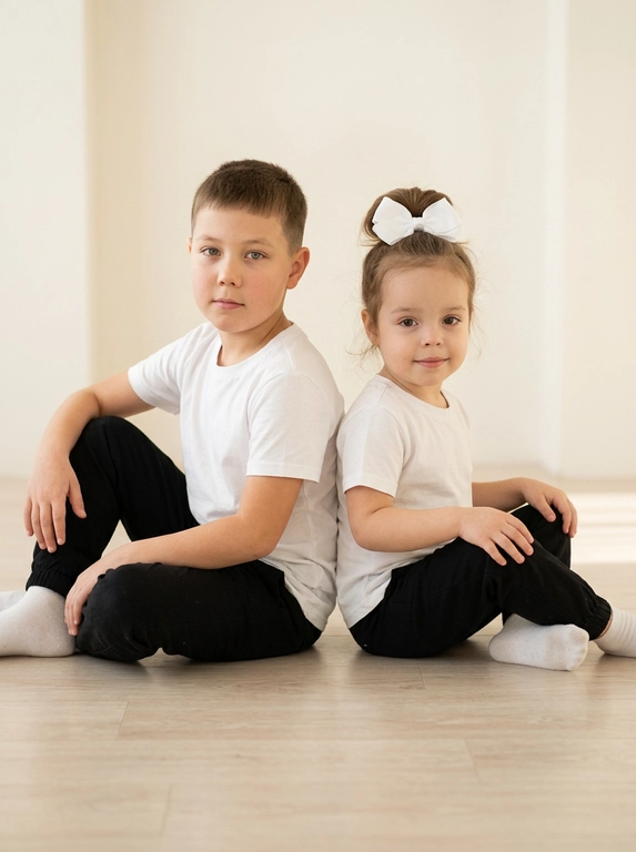 Сохранив черты лица детей изобразить мальчика и девочку сидящими на полу рядом с друг другом спина к спине. Они смотрят в камеру. Одежда белые футболки и черные легкие штаны, белые носки. Фон студии в светлых тонах. У девочки собраны волосы сверху на голове и завязаны белым бантом. Возраст девочки 3 года. Возраст мальчика 9 лет