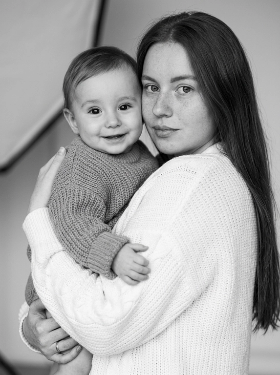 Создай фотореалистичный черно-белый студийный портрет, не меняя черты лица. Эстетичная модель нежно держит на руках ребенка. Модель повернута спиной к камере, слегка разворачивает голову через плечо и смотрит прямо в объектив спокойным, теплым взглядом. На ней свободный белый вязаный кардиган с мягкой объемной фактурой, естественно ниспадающий по плечам. Ребенок одет в объемный вязаный фактурный свитер, нежно прижимается к модели и искренне улыбается, глядя в камеру. Композиция тесная и эмоциональная, кадр построен так, чтобы подчеркнуть ощущение близости и тепла между персонажами.

Мягкий направленный студийный свет с большим рассеивателем (в кадре нет оборудования - студийного света с рассеивателем) создает плавные переходы света и тени, глубокий объем и выразительный светотеневой рисунок. Свет красиво подчеркивает естественную текстуру кожи, объем ткани и форму лица. Фон чистый, минималистичный и слегка размытый, без отвлекающих деталей.

Fine art portrait photography, luxury editorial style, intimate family portrait, elegant minimalist composition, medium close-up, eye-level camera, shallow depth of field, soft focus, realistic film grain, monochrome masterpiece, rich grayscale tonal range, cinematic contrast, natural skin texture, visible pores, highly detailed knitted fabric, ultra photorealistic, 85mm lens, f/2, HDR, extremely realistic, museum-quality portrait, no AI look.
