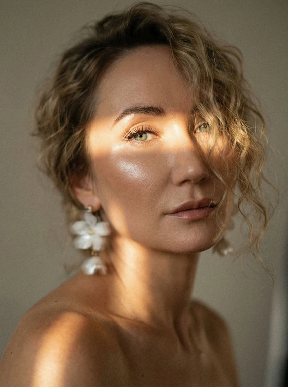 Кинематографический эффект с добавлением зерна. Ультрареалистичный, экстремально крупный портрет части лица девушки: на неё падает солнечный свет полосой, освещая глаза; её голова немного повёрнута в сторону. Тело подсвечено хайлайтером. Макияж лёгкий, с насыщенным хайлайтером; ресницы длинные, пышные. Она смотрит в камеру со спокойным, созерцательным выражением. Освещение мягкое, с лёгким акцентом на её лицо, создаёт нежные тени и подчёркивает черты. В ушах — серьги в виде белых цветов. Фон минималистичный и нейтральный, что усиливает спокойную и элегантную атмосферу. Фото смазанное, как будто сделано в движении, с двойной экспозицией.


Какая будет прическа?: Волосы кудрявые, частично падают на лицо.