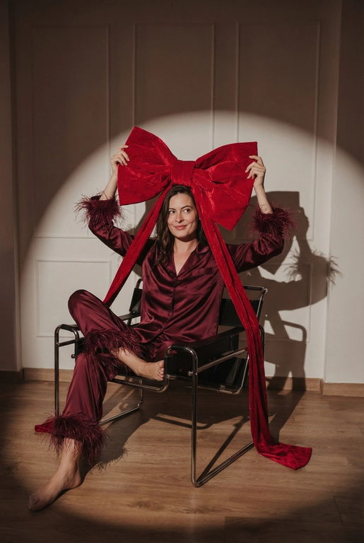 Создай фэшн-фото человека крупным планом до колен, позирующего на стуле под направленным светом прожектора на фоне стены с лепниной, как на референсном изображении. Черты лица, пол, комплекция, возраст и цвет волос должны быть такими же, как на референсе. Драматичное сценическое освещение с выраженным световым пятном и мягкими тенями. Человек сидит уверенно и расслабленно. Одна нога на сидении,  другая на полу. В руках человек держит очень большой красный бархатный бант над головой — жест выглядит игриво и символично, создавая ощущение, что человек сам является подарком. У банта длинные ленты («ножки»), которые свободно и естественно ниспадают вниз. Взгляд направлен немного в сторону, мягкая улыбка. Общая атмосфера: high-fashion, новогодний гламур, рождественское настроение. Стиль журнальной съёмки, кинематографичный свет, высокая детализация, чистая композиция, профессиональная студийная фотография. На человеке шелковая пижама глубокого бордового цвета: свободная рубашка и брюки. Манжеты рукавов и края брюк отделаны страусиным пером. Ткань с мягким благородным блеском, подчёркивающим фактуру шёлка.