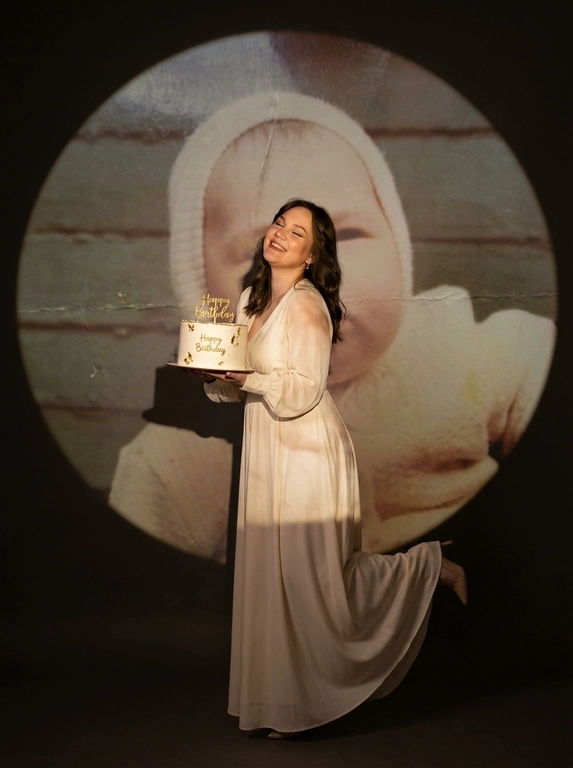 Create ultra-realistic cinematic birthday photoshoot, elegant studio with a dark background and a large circular spotlight projection on the wall. Inside the spotlight, a faded vintage childhood photo of the same girl(the reference image 2nd given)appears as a nostalgic memory, slightly transparent with old photo texture and soft blur. In the foreground, the present-day version of the same model (the reference image 1st given)  holding a white birthday cake decorated with gold accents with a “Happy Birthday". 

If the model is a woman, then she is wearing a cream long gown. If the model is a man, he is wearing a suit.

her head tilted back, eyes closed, natural smile,  cinematic shadow cast on the wall, warm soft spotlight lighting.

If the model was a woman, she additionally lifted her leg in an elegant pose.

Доп. характеристики: Фигура: средней полноты, волосы, чуть ниже плеч