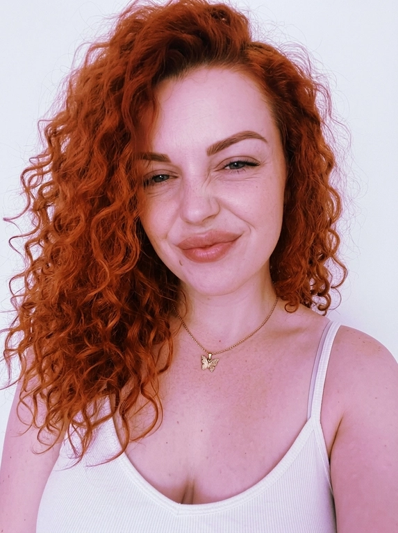 Vertical close-up portrait in the style of a natural selfie: a woman with her head tilted and a soft playful expression, one eye squinted or closed, gaze directed at the camera.
Expressive full lips, neat eyebrows, and soft makeup with an emphasis on the lips and eyes are noticeable on the face. There are thin chains around her neck, one with a small butterfly pendant.
The clothes are a white ribbed tank top with thin straps, revealing the shoulders and upper chest. The background is almost completely white, empty and minimalistic, like a light wall, without unnecessary objects.
The light is very soft, diffused, slightly overexposed, with a cool pink and purple hue, creating a gentle, slightly dreamy atmosphere.
The composition is close and intimate, the frame is cropped around the shoulders and chest, with emphasis on the face. The camera is at face level, creating the feeling of taking a picture from a phone. Light graininess, a gentle loss of detail in the highlights, natural, fashionable, slightly vintage aesthetic.



Причёска: Не менять прическу