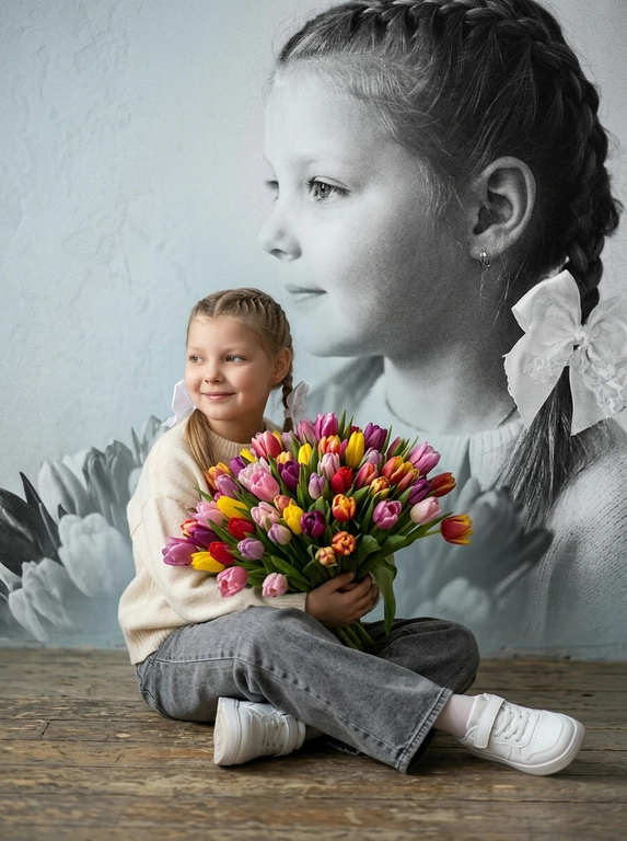 Создай портрет, не меняя черты лица, и превратите его в художественный портрет.
Ребенок сидит на полу в изящной позе, в белой кофте, мягких свободных серых джинсах и белых спортивных кроссовках. Ребенок держит в руках огромный букет ярких разноцветных тюльпанов.
Выражение лица — лёгкая улыбка, ребенок смотрит в сторону. 
Фон — тот же его профиль, в мягком крупном чёрно-белом художественном стиле, крупный план сбоку.
Композиция должна быть художественной, мягкой, реалистичной.
Создай изображение, которое выглядит как настоящая фотография. 
Не меняй черты лица.