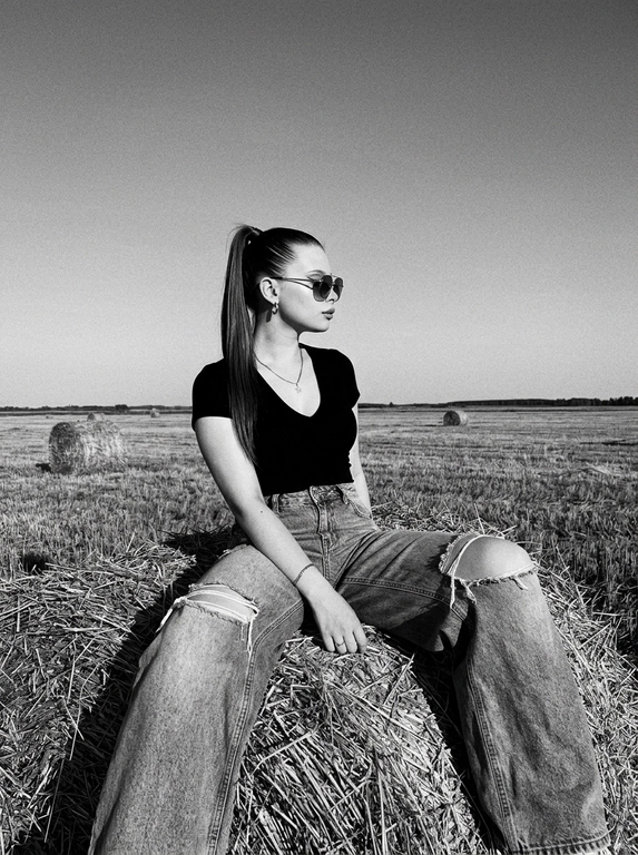 Чёрно-белая high-fashion editorial фотография в сельской местности, женщина с внешностью как на референсе, черты лица не изменять, композиция — более общий план (wide shot), камера на среднем расстоянии, фигура полностью в кадре с окружающим пространством, она сидит на большом тюке сена в поле, корпус слегка развернут, одна нога свободно опущена вниз, вторая согнута, создавая расслабленную, но выразительную позу, руки естественно лежат на колене и на поверхности тюка, взгляд направлен в сторону, профиль, выражение спокойное и уверенное, одежда: чёрный топ с V-образным вырезом и объёмные светлые широкие джинсы с потёртостями и рваными элементами, аксессуары: крупные солнцезащитные очки, минималистичные украшения, фон: открытое поле с несколькими тюками сена на разной глубине, создающими перспективу, линия горизонта ниже центра кадра, чтобы подчеркнуть небо, освещение: жёсткий естественный дневной свет с мягкими облаками, формирует выразительные тени и контраст, стиль: кинематографический, плёночное зерно, высокая детализация, реалистичная текстура кожи и ткани, редакционный fashion стиль, акцент на атмосфере и пространстве кадра


Какая будет прическа?: Гладкие волосы собраны в высокий хвост