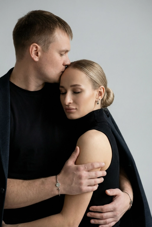   Мужчина и женщина позируют в студии в объятиях. Мужчина стоит позади женщины, обнимая её обеими руками; его тело повернуто к ней, голова слегка наклонена вниз, губы касаются её лба. Женщина немного отклоняет голову назад к его плечу, её глаза закрыты. Кадр — средний, от талии до головы, снят на уровне глаз. Композиция плотная, акцентирует линии рук и соприкосновение лиц. Женщина стоит, прямо обращённая к камере.
На ней облегающий чёрный топ без рукавов с высоким воротом, из плотной матовой ткани.
Мужчина одет в чёрную футболку и чёрную неброскую куртку. На обоих запястьях — тонкие металлические браслеты с минималистичными круглыми подвесками, а женщина носит небольшие светлые металлические серьги с гладкой поверхностью.
Обстановка минималистичная: однотонный светло-серый фон без текстуры.
Освещение — мягкий студийный свет: рассеянный фронтальный и боковой свет создаёт плавные переходы света и тени на лицах и руках. Тени мягкие, средней глубины. Контраст умеренный, без жёстких бликов. Свет равномерно распределён по фигурам, фон подсвечен мягко, без выраженных градиентов.
Съёмка на портретный объектив 85 мм, малая глубина резкости с чётким фокусом на лицах и руках; фон слегка размыт, текстуры кожи и ткани высоко детализированы. Это минималистичный студийный портрет, высокодетализированный и фотореалистичный.
Минималистичное романтическое студийное настроение, нейтральные тёплые тона кожи, низкая–средняя насыщенность, мягкое рассеянное освещение, средний контраст с нежными тенями, плавные переходы бликов, естественная передача кожи, чистый светлый фон, тонкий тональный баланс, современная мягкая редакционная цветокоррекция. Волосы женщины собраны в низкий, гладкий пучок с аккуратно уложенными прядями.