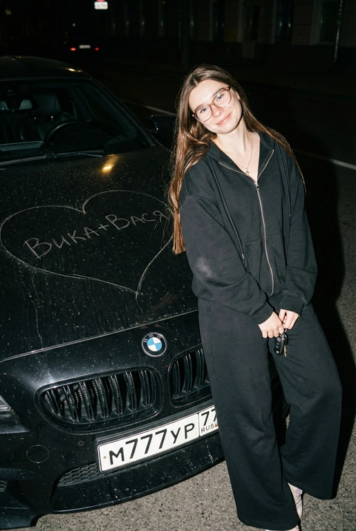 Строго сохранить лицо с прикреплённой референс-фотографии, на 100% без искажений. Человек одет полностью в чёрный, монохромный оверсайз спортивный костюм: свободная худи на молнии и широкие расслабленные штаны, без логотипов, текста и брендинга. Акцент на реальной текстуре кожи и детализированной текстуре ткани.
Сырой кинематографичный ночной кадр. Человек опирается на капот автомобиля, средний план, лёгкий верхний ракурс. В одной руке игриво держит ключи от машины. Номерной знак виден: m777yp 77.
Строго по центру капота, покрывая всю поверхность капота, глубоко и грубо процарапаны (не мелом) царапины: большое сердце с текстом «» внутри.
Жёсткая прямая вспышка, сильные тени, высокий контраст, видимая пыль и микро-царапины на краске. Тёмный размытый городской фон.
Объектив 35mm, f/2.8, грубая уличная эстетика.

Надпись на капоте: Вика+Вася

Марка автомобиля: Опирается на чёрный BMW M6 F90