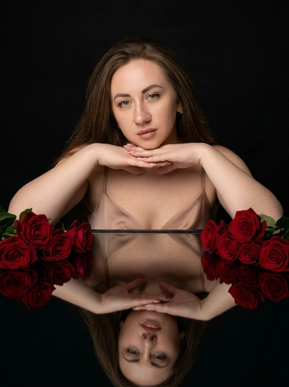 Студийный портрет женщины. Обстановка минималистичная: черный, глянцевый стол, отражающий человека и объекты на нем. Стиль фото - гламурная съемка с акцентом на красоту. Атмосфера спокойная и немного загадочная. Цветовая гамма - сочетание глубокого черного фона, красных роз и телесных оттенков кожи.

Женщина лежит на столе, опираясь на предплечья. Руки сложены, как будто она придерживает голову. На ней надет топ или платье с тонкими бретельками телесного цвета. На столе, вдоль рук человека, лежат красные розы. Они расположены таким образом, чтобы создавать симметричную композицию. Отражение в столе дублирует изображение человека и роз, усиливая эффект глубины и симметрии. Свет мягкий, рассеянный, акцентирует внимание на лице и розах, создавая игру света и теней.