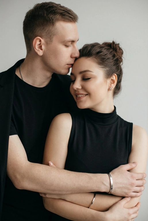 Мужчина и женщина позируют в студии в объятиях. Мужчина стоит позади женщины, обнимая её обеими руками; его тело повернуто к ней, голова слегка наклонена вниз, губы касаются её лба. Женщина немного отклоняет голову назад к его плечу, её глаза закрыты. Кадр — средний, от талии до головы, снят на уровне глаз. Композиция плотная, акцентирует линии рук и соприкосновение лиц. Женщина стоит, прямо обращённая к камере.
На ней облегающий чёрный топ без рукавов с высоким воротом, из плотной матовой ткани.
Мужчина одет в чёрную футболку и чёрную неброскую куртку. На обоих запястьях — тонкие металлические браслеты с минималистичными круглыми подвесками, а женщина носит небольшие светлые металлические серьги с гладкой поверхностью.
Обстановка минималистичная: однотонный светло-серый фон без текстуры.
Освещение — мягкий студийный свет: рассеянный фронтальный и боковой свет создаёт плавные переходы света и тени на лицах и руках. Тени мягкие, средней глубины. Контраст умеренный, без жёстких бликов. Свет равномерно распределён по фигурам, фон подсвечен мягко, без выраженных градиентов.
Съёмка на портретный объектив 85 мм, малая глубина резкости с чётким фокусом на лицах и руках; фон слегка размыт, текстуры кожи и ткани высоко детализированы. Это минималистичный студийный портрет, высокодетализированный и фотореалистичный.
Минималистичное романтическое студийное настроение, нейтральные тёплые тона кожи, низкая–средняя насыщенность, мягкое рассеянное освещение, средний контраст с нежными тенями, плавные переходы бликов, естественная передача кожи, чистый светлый фон, тонкий тональный баланс, современная мягкая редакционная цветокоррекция.

Не изменять мою прическу