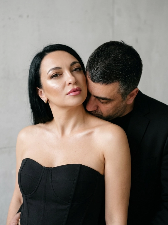 Не меняя черты лица, студийная fashion-съемка пары в минималистичном интерьере с серо-белым фоном, атмосфера гламура и интимной близости, ракурс — крупный портрет по грудь с легким поворотом корпуса, женщина на переднем плане, мужчина позади склоняется к её плечу, поза — она слегка отвернута от камеры с прямым взглядом, он касается лицом её шеи/плеча, одежда — на женщине черный корсет/бандо с открытыми плечами, на мужчине классический черный пиджак и рубашка, макияж — glam (ровный сияющий тон, сильный хайлайтер, четкий контуринг, стрелки, густые ресницы, глянцевые губы), свет — жесткая фронтальная вспышка + мягкий заполняющий свет, выраженные тени позади, эффект flash photography / editorial, холодно-нейтральная цветокоррекция с контрастом и глянцем кожи, высокая детализация, slight film grain, shallow depth of field, съемка на Canon EOS R / 50mm, f/2.0, direct flash, studio look.