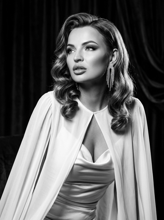 Фотореалистичный контрастный чёрно-белый glamour/editorial портрет в стиле Old Hollywood, вертикальный кадр по грудь. Женщина в 3/4, корпус чуть наклонён вперёд; голова повернута влево, взгляд вверх/в сторону (не в камеру), губы чуть приоткрыты, выражение драматичное и кинематографичное. Образ: белая накидка из шифона с длинными струящимися рукавами, под накидкой драпированный атласный белый топ; на ушах крупные серьги со стразами/каскадом. Макияж: классический вечерний glam (в ч/б читается как контраст) — чёткие брови, мягкие smoky тени, тонкая стрелка, длинные ресницы, губы с чётким контуром и сатиновым финишем. Фон: тёмный студийный с мягким рисунком/драпировкой (как бархатные шторы), размытый. Свет: жёсткий key слева спереди (beauty dish) + контровой/rim справа для отделения волос, глубокие тени, яркие блики на скулах и волосах, богатый тональный диапазон (глубокие чёрные и сливочные белые). Камера: 85mm, f/2.0–2.8, ISO 100–200, 1/160, резкость на глазах и губах, фон в мягком боке, лёгкое плёночное зерно, без “пластика”.

Какая будет укладка? : Волосы: объёмные голливудские волны, аккуратная укладка с сильным объёмом и мягкими завитками, блеск волос.