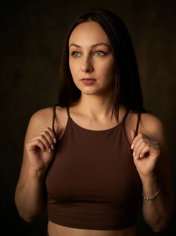 Максимально реалистичная фотография в стиле moody boudoir editorial, передающая атмосферу интимности, чувственности и приглушенного драматизма.
Крупный план по пояс (waist-up shot / close-up). женщина позирует в полумраке, придерживая руками края одежды. [Поза/взгляд/эмоция]: Плечи слегка приподняты, руки с длинным светлым маникюром сжимают тонкие бретели черного топа; голова немного повернута в сторону, взгляд задумчивый и отведен в сторону; выражение лица томное, загадочное и уверенное.
Вечерний «glam» выразительные стрелки, густо накрашенные ресницы, создающие эффект «кошачьего взгляда», мягкий контуринг лица и матовая помада цвета пыльной розы.
Облегающий черный кроп-топ на очень тонких бретелях-спагетти. Материал — матовая эластичная ткань. На запястье виден изящный серебряный браслет.
Минималистичный интерьер в темных тонах. Фон однотонный, слегка текстурированный, уходящий в глубокую тень, что создает эффект камерности.
 Низкий ключ (low-key lighting). Мягкий, теплый направленный свет падает сбоку, подчеркивая рельеф ключиц, линию плеч и текстуру кожи. Глубокие, кинематографичные тени создают объем и драматичное настроение.
 85mm f/1.4, идеальное фокусное расстояние для портрета, создающее минимальные искажения и сильное размытие фона. Очень малая глубина резкости; фокус идеально наведен на глаза и лицо модели, в то время как плечи и руки уходят в мягкое расфокусирование.
Гиперреалистичная текстура кожи: видны поры, естественный блеск на выступающих частях лица и плеч. Отсутствие «пластикового» эффекта ретуши.
 текстура ткани топа под пальцами, тонкие волоски на макушке, мягкий градиент тени на стене.
Эстетика «dark feminine energy», интимная, теплая и дорогая атмосфера (expensive moody vibe).

Какая будет прическа?: Не изменять мою прическу

Доп. характеристики: Топ закрытый, шоколадного цвета, глаза зеленые,легкий макияж на лице, лицо не менять