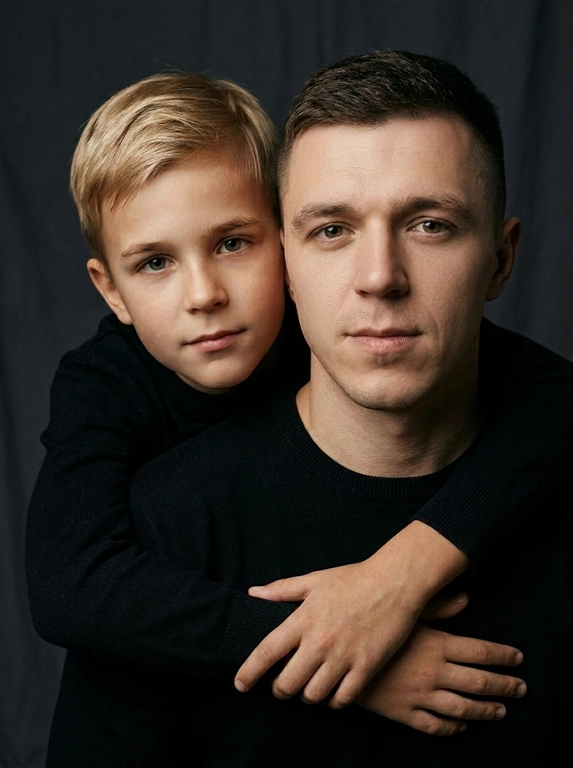 Создай портретный студийный кадр из фотосессии отца и сына. Сын обнимает отца сзади, лежа подбородком на его плече, руки мягко сомкнуты на груди мужчины. Оба смотрят прямо в камеру. Естественные спокойные эмоции, лёгкая серьёзность во взгляде.
Одежда — однотонные чёрные лонгсливы или свитера без принтов. Тёмный нейтральный фон (глубокий серо-оливковый или графитовый). Мягкий направленный студийный свет с лёгкой теневой глубиной, акцент на лицах, тёплая естественная цветокоррекция, высокая детализация кожи, натуральная текстура без излишней ретуши.
Кадр — крупный или средний план, плотная композиция, лица расположены близко друг к другу. Атмосфера — тёплая, камерная, эмоциональная, ощущение близости и доверия. Профессиональная съёмка, 85mm lens, shallow depth of field, cinematic portrait, ultra realistic.