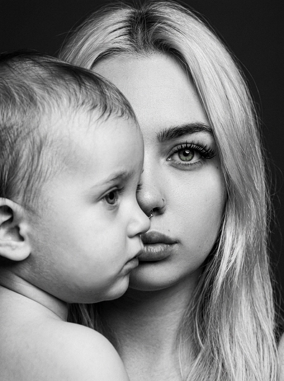Чёрно-белый фотореалистичный студийный beauty-fashion портрет родителя и ребёнка с представленных фото. Крупный плотный кадр по плечи, без свободного пространства. Ребёнок находится на переднем плане, ближе к камере, в профиль, расположен сбоку и частично перекрывает лицо родителя. Лицо ребёнка закрывает примерно половину лица родителя, создавая сильный эффект визуального перекрытия. Родитель находится позади ребёнка, строго по центру кадра; у родителя виден только один глаз, направленный прямо в камеру, вторая часть лица скрыта лицом ребёнка. Лица расположены максимально близко, почти соприкасаются, но без явного физического действия. Ребёнок смотрит в сторону, выражение спокойное, сдержанное и закрытое. Родитель смотрит прямо в камеру одним открытым глазом, взгляд холодный, спокойный и пронизывающий. Атмосфера high-fashion editorial: минимальная эмоциональность, люксовая отстранённость, скрытое напряжение и холодная близость. Фон тёмный, чистый, студийный, без деталей. Жёсткое студийное освещение, высокий контраст, глубокие тени, выразительная светотень на лицах, натуральная текстура кожи, sharp focus, dramatic black and white photography, editorial beauty portrait, без улыбок, без лишних предметов, без текста и логотипов.