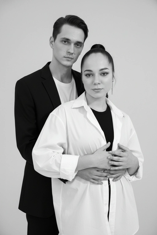 Черно-белый портрет мужчины и женщины в стиле фэшн фотосессии в фотостудии. Не изменяйте лица. Фон нейтральный, однотонный, светло-серый. Атмосфера интимная и спокойная. На переднем плане женщина. Вся композиция выполнена в приглушенной монохромной гамме со студийным освещением, которое подчеркивает текстуру кожи и одежды. Её руки лежат на груди мужчины. Мужчина обнимает женщину сзади, положив свою руку поверх её руки. Она одета в черный топ, белую оверсайз рубашку. Мужчина одет в  классическом черном пиджаке и белой футболке.