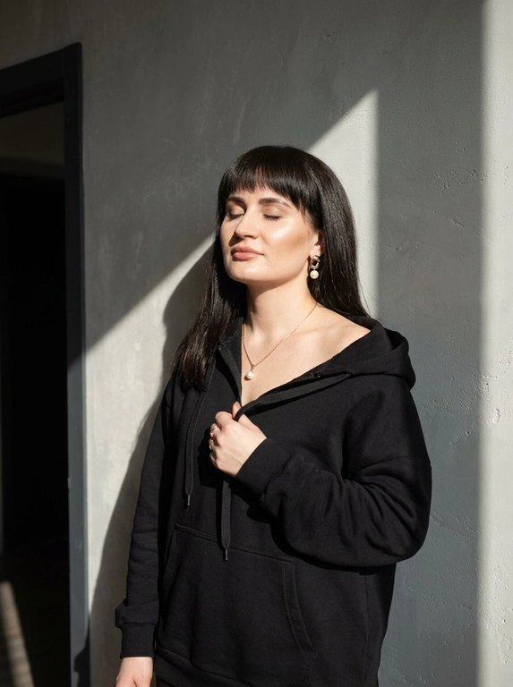 Используй загруженное фото для лица, строго не меняя черты лица, пропорции, возраст, текстуру кожи и волосы. Минималистичный портрет в интерьере с естественным светом, женщина стоит у серой текстурированной оштукатуренной стены, голова слегка запрокинута назад, глаза закрыты, лицо повернуто к источнику света, правая рука держит ткань толстовки у груди, левая рука расслабленно опущена вдоль тела. Оверсайз толстовка с капюшоном спущена с правого плеча, видны завязки, на шее тонкая золотая цепочка с кулоном с жемчужиной, золотые серьги среднего размера с жемчугом, золотое кольцо на пальце. Естественный макияж с деликатным скульптурированием, влажное свечение кожи, кожа ультрареалистичная без эффекта пластика и чрезмерной ретуши. Жёсткий направленный солнечный свет из окна создаёт резкие графичные тени на стене и лице, контрастное освещение, слева тёмный дверной проём добавляет глубину пространству. Съёмка на 50mm f/2.0, малая глубина резкости, естественный оконный свет, выраженные тени, цветовая гамма бежевый, золото, серый, баланс тёплых и холодных оттенков, minimalist editorial, фотореализм высокого качества.

Какой цвет толстовки?: Цвет толстовки чёрный.