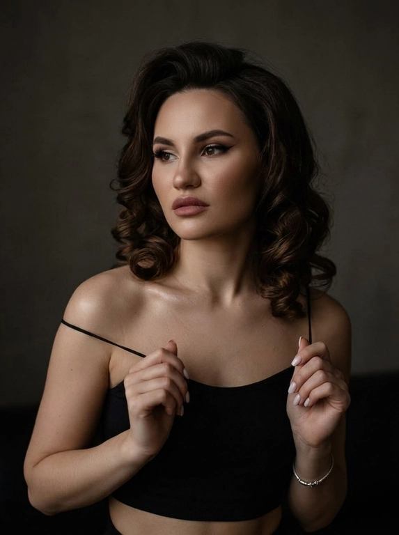 Максимально реалистичная фотография в стиле moody boudoir editorial, передающая атмосферу интимности, чувственности и приглушенного драматизма.
Крупный план по пояс (waist-up shot / close-up). женщина позирует в полумраке, придерживая руками края одежды. [Поза/взгляд/эмоция]: Плечи слегка приподняты, руки с длинным светлым маникюром сжимают тонкие бретели черного топа; голова немного повернута в сторону, взгляд задумчивый и отведен в сторону; выражение лица томное, загадочное и уверенное.
Вечерний «glam» выразительные стрелки, густо накрашенные ресницы, создающие эффект «кошачьего взгляда», мягкий контуринг лица и матовая помада цвета пыльной розы.
Облегающий черный кроп-топ на очень тонких бретелях-спагетти. Материал — матовая эластичная ткань. На запястье виден изящный серебряный браслет.
Минималистичный интерьер в темных тонах. Фон однотонный, слегка текстурированный, уходящий в глубокую тень, что создает эффект камерности.
 Низкий ключ (low-key lighting). Мягкий, теплый направленный свет падает сбоку, подчеркивая рельеф ключиц, линию плеч и текстуру кожи. Глубокие, кинематографичные тени создают объем и драматичное настроение.
 85mm f/1.4, идеальное фокусное расстояние для портрета, создающее минимальные искажения и сильное размытие фона. Очень малая глубина резкости; фокус идеально наведен на глаза и лицо модели, в то время как плечи и руки уходят в мягкое расфокусирование.
Гиперреалистичная текстура кожи: видны поры, естественный блеск на выступающих частях лица и плеч. Отсутствие «пластикового» эффекта ретуши.
 текстура ткани топа под пальцами, тонкие волоски на макушке, мягкий градиент тени на стене.
Эстетика «dark feminine energy», интимная, теплая и дорогая атмосфера (expensive moody vibe).

Какая будет прическа?: Уложенные в крупные классические локоны с заметным объемом у корней. Прическа выглядит аккуратной и глянцевой.