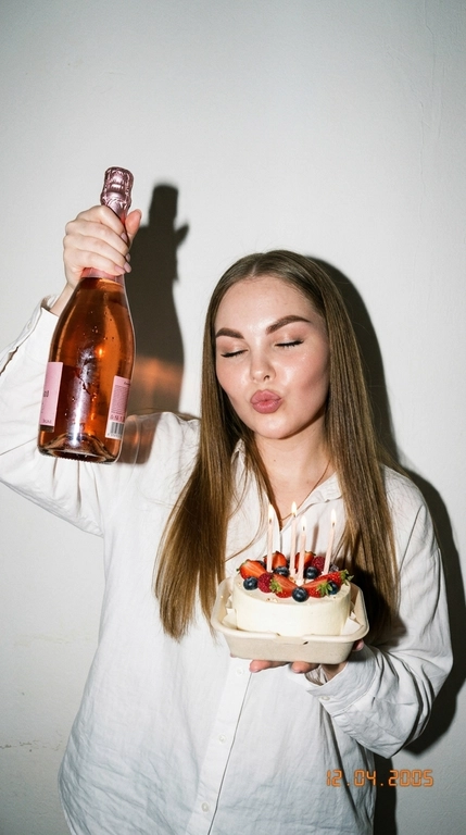 Ультрареалистичная фотография в эстетике Party Girl / Birthday Aesthetic, стилизация под случайный снимок на компактную цифровую камеру начала 2000-х.
 девушка празднует день рождения. На ней свободная белая рубашка оверсайз на пуговицах. В одной руке она высоко держит бутылку розового шампанского, в другой — небольшой бенто-торт со светлыми свечами и свежими ягодами.

Глаза закрыты, на лице игривое выражение с лёгким «поцелуйным» жестом губ. Не меняй прическу модели.

Съёмка со вспышкой прямо в камеру на фоне простой молочно-белой стены. Жёсткий свет создаёт характерную чёткую тень за фигурой, яркие блики на коже, волосах, бутылке и глазури торта. Атмосфера спонтанного домашнего праздничного снимка: немного несовершенная композиция, лёгкая зернистость, естественная текстура кожи, реалистичные отражения и мелкие детали.

Стиль: 2000s digital camera, lo-fi party aesthetic, candid snapshot, hard flash, authentic lifestyle photography, natural skin texture, realistic colors, high detail, 4K.

Без: студийной постановки, мягкого идеального света, пластиковой кожи, чрезмерной ретуши, CGI, мультяшности, искажённых рук и пальцев, лишних пальцев, деформированной бутылки или торта, артефактов текста, логотипов и водяных знаков.