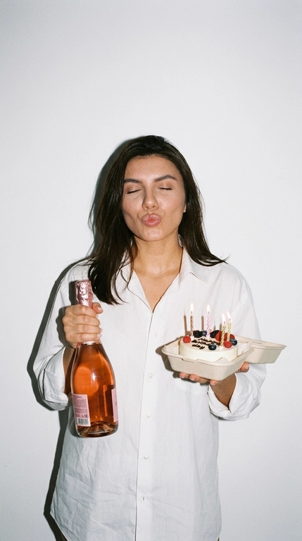 Ультрареалистичная фотография в эстетике Party Girl / Birthday Aesthetic, стилизация под случайный снимок на компактную цифровую камеру начала 2000-х.
 девушка празднует день рождения. На ней свободная белая рубашка оверсайз на пуговицах. В одной руке она высоко держит бутылку розового шампанского, в другой — небольшой бенто-торт со светлыми свечами и свежими ягодами.

Глаза закрыты, на лице игривое выражение с лёгким «поцелуйным» жестом губ. Не меняй прическу модели.

Съёмка со вспышкой прямо в камеру на фоне простой молочно-белой стены. Жёсткий свет создаёт характерную чёткую тень за фигурой, яркие блики на коже, волосах, бутылке и глазури торта. Атмосфера спонтанного домашнего праздничного снимка: немного несовершенная композиция, лёгкая зернистость, естественная текстура кожи, реалистичные отражения и мелкие детали.

Стиль: 2000s digital camera, lo-fi party aesthetic, candid snapshot, hard flash, authentic lifestyle photography, natural skin texture, realistic colors, high detail, 4K.

Без: студийной постановки, мягкого идеального света, пластиковой кожи, чрезмерной ретуши, CGI, мультяшности, искажённых рук и пальцев, лишних пальцев, деформированной бутылки или торта, артефактов текста, логотипов и водяных знаков.