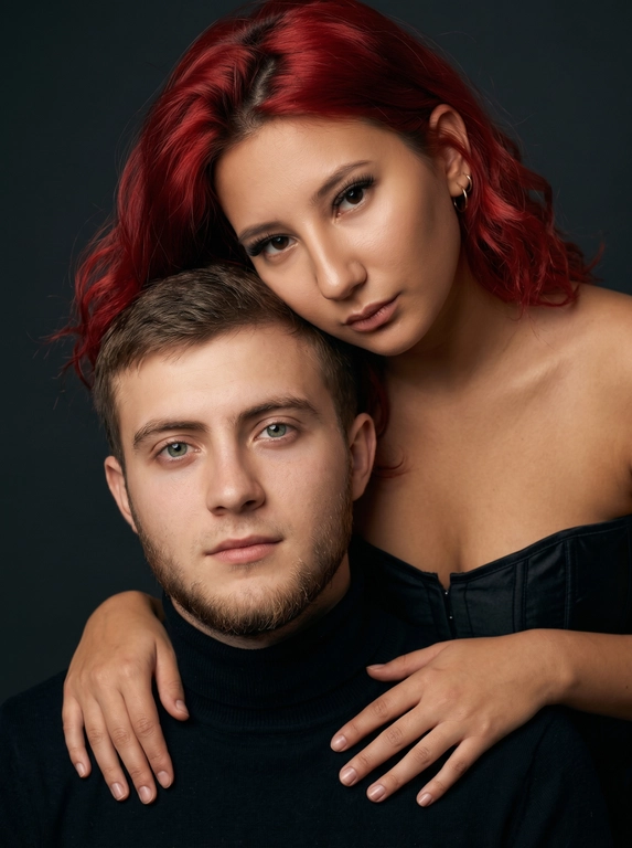 Гиперреалистичный студийный fashion-портрет молодой пары по загруженному референсу, 100% сходство лиц. Черты лица не изменять. Крупный план, кадр по плечи. Мужчина, спокойный уверенный взгляд прямо в камеру, естественное выражение лица, одет в черный трикотажный свитер с высоким воротом. Женщина стоит позади мужчины и нежно обнимает его, ее руки лежат на его плечах. Ее лицо расположено рядом с его лицом, она повернута слегка боком, голова немного наклонена вперед, взгляд направлен прямо в камеру. У женщины выразительные глаза с длинными густыми черными ресницами, естественный clean-glam макияж, натуральные брови, теплый оливковый оттенок кожи, небольшие серьги-кольца. Композиция плотная и интимная, их лица расположены очень близко друг к другу, современный модный портрет пары. Женщина одета в черный корсет. Кожа максимально натуральная: живая текстура кожи, видимые микропоры, subtle skin glow, естественный мягкий блеск кожи, natural skin microtexture, skin translucency, micro skin details, легкие естественные несовершенства кожи, без ретуши и без эффекта пластика. Освещение: мягкий студийный beauty lighting, рассеянный ключевой свет спереди и немного сбоку, мягкие глубокие тени для объема лица, деликатные световые блики на коже. Фон: глубокий темный студийный фон, темно-графитовый или почти черный, создающий контраст с кожей и лицами. Стиль: luxury fashion editorial photography, high fashion portrait, ultra photorealistic, 85mm portrait lens, shallow depth of field, creamy bokeh, cinematic soft lighting, RAW photo, extremely detailed, fine hair strands, natural skin pores, professional photography, photorealism, 8k, high dynamic range.