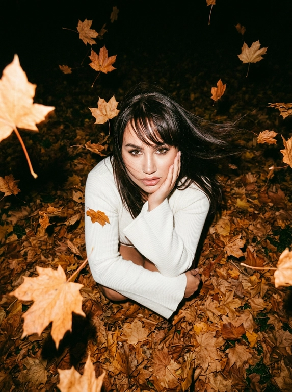Ultra-realistic candid NIGHT autumn photo. The woman sits curled up directly in a thick carpet of dry autumn maple leaves, knees pulled toward her chest. Camera positioned very high above her, almost directly overhead, dramatic top-down perspective. She raises her face toward the camera, one hand gently supporting her cheek, the other arm resting across her knees.
She wears a fitted white knitted top with very long dramatic FLARED BELL SLEEVES, fitted through the arms and strongly widening from the elbows.
Strong autumn wind blows her hair.

It is DARK outside, almost nighttime. The background beyond the flash falls into deep natural darkness. Shot with a powerful direct on-camera flash: her face, white sweater and the closest leaves are brightly illuminated, while distant leaves rapidly disappear into black shadows. Dozens of dry orange, rust and brown leaves are FLYING THROUGH THE AIR around her, caught mid-flight by the flash; some pass very close to the lens and create strong realistic motion blur and streaks. A few strands of hair also have motion blur from the wind. Face remains sharp. Raw spontaneous flash photography, high contrast, deep blacks, warm saturated autumn tones, slight grain, imperfect handheld framing, realistic textures, no studio lighting, no HDR, no AI look.