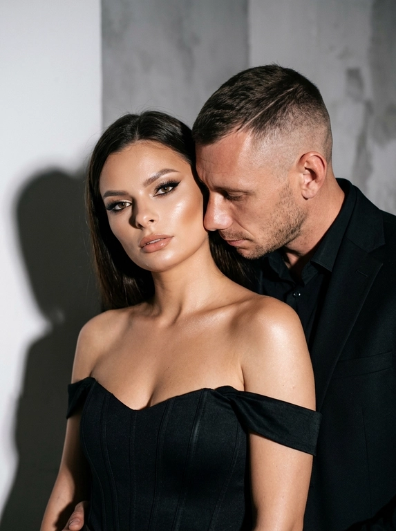 Не меняя черты лица, студийная fashion-съемка пары в минималистичном интерьере с серо-белым фоном, атмосфера гламура и интимной близости, ракурс — крупный портрет по грудь с легким поворотом корпуса, женщина на переднем плане, мужчина позади склоняется к её плечу, поза — она слегка отвернута от камеры с прямым взглядом, он касается лицом её шеи/плеча, одежда — на женщине черный корсет/бандо с открытыми плечами, на мужчине классический черный пиджак и рубашка, макияж — glam (ровный сияющий тон, сильный хайлайтер, четкий контуринг, стрелки, густые ресницы, глянцевые губы), свет — жесткая фронтальная вспышка + мягкий заполняющий свет, выраженные тени позади, эффект flash photography / editorial, холодно-нейтральная цветокоррекция с контрастом и глянцем кожи, высокая детализация, slight film grain, shallow depth of field, съемка на Canon EOS R / 50mm, f/2.0, direct flash, studio look.