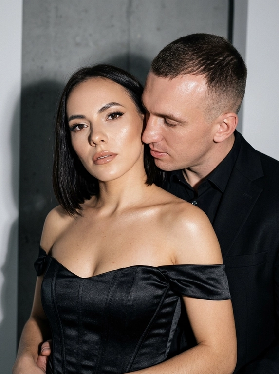 Не меняя черты лица, студийная fashion-съемка пары в минималистичном интерьере с серо-белым фоном, атмосфера гламура и интимной близости, ракурс — крупный портрет по грудь с легким поворотом корпуса, женщина на переднем плане, мужчина позади склоняется к её плечу, поза — она слегка отвернута от камеры с прямым взглядом, он касается лицом её шеи/плеча, одежда — на женщине черный корсет/бандо с открытыми плечами, на мужчине классический черный пиджак и рубашка, макияж — glam (ровный сияющий тон, сильный хайлайтер, четкий контуринг, стрелки, густые ресницы, глянцевые губы), свет — жесткая фронтальная вспышка + мягкий заполняющий свет, выраженные тени позади, эффект flash photography / editorial, холодно-нейтральная цветокоррекция с контрастом и глянцем кожи, высокая детализация, slight film grain, shallow depth of field, съемка на Canon EOS R / 50mm, f/2.0, direct flash, studio look.